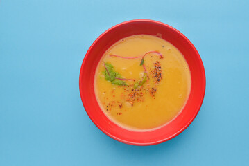 Squash soup with rosemary and paprika. cream soup served in a red bowl over pastel blue background.