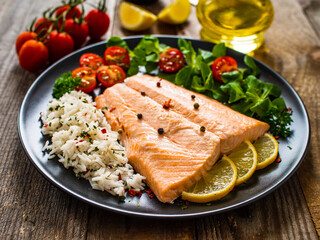 Fried salmon fillet with fried potatoes, lemon and vegetable salad served on black plate on wooden table
