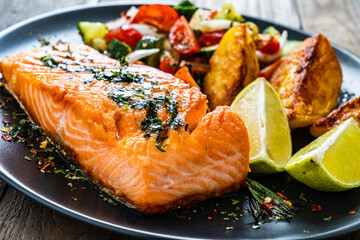 Fried salmon fillet with fried potatoes, lime and vegetable salad served on black plate on wooden table
