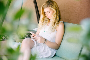 Young blond haired woman texting on her phone.  She is outdoors in a southwestern beige and light...