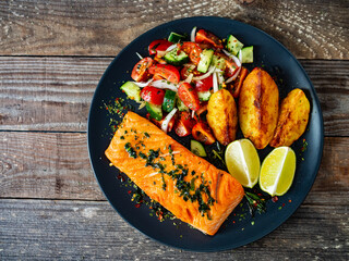 Fried salmon fillet with fried potatoes, lime and vegetable salad served on black plate on wooden...