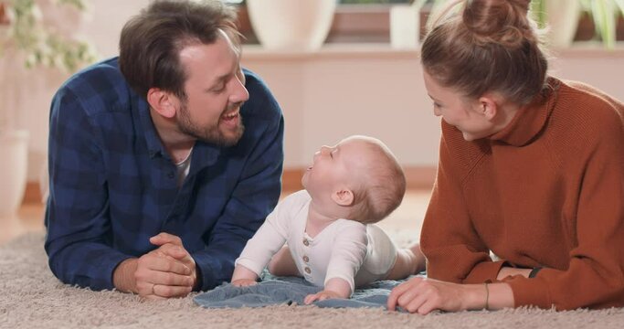 Smiling young couple lying together on rug on their living room floor at home with their adorable baby. Baby smiles and looks at the bearded father, who kisses it, mother looks at them.