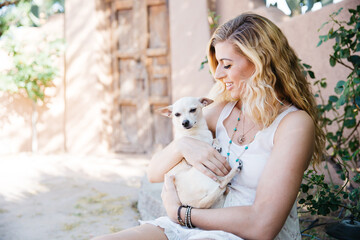 Young blond haired woman holding cuddling a blond short haired Chihuahua dog in her arms with loving affection.  She is outdoors in a southwestern beige and light green environment.  She is wearing an