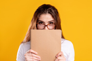Teacher portrait. Business coaching. Confident smart woman in glasses hiding behind beige blank notebook isolated on orange copy space background. Professional motivation. Knowledge education.