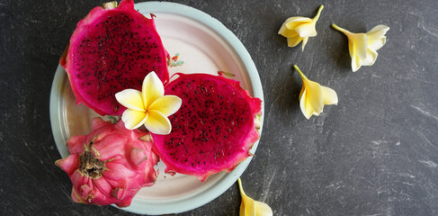 Obraz na płótnie Canvas two slices of red dragon fruit and one whole dragon fruit, served on a tray and decorated with frangipani flowers, on a stone pad