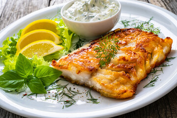 Fish dish - fried cod fillet with cream sauce and lemon served on wooden table
