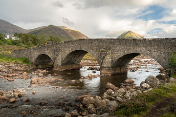 Fototapeta na wymiar Old stone arch bridge over a mountain river at Sligachan on the Isle of Skye in the Highlands of Scotland, the Cuillin mountains rising behind lit by sunset