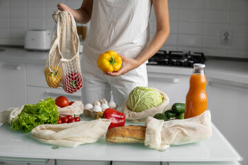 Woman hand holding Reusable eco textile grocery bag with vegetables and takes red and yellow pepper out. Zero waste and plastic free concept. Girl is holding mesh cotton shopper with vegetables.