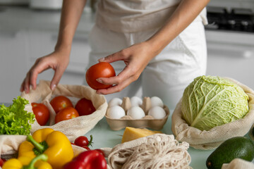 Woman hand holding Reusable eco textile grocery bag with vegetables and takes red tomato out. Zero waste and plastic free concept. Girl is holding mesh cotton shopper with vegetables.