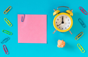 Blue background, pink card with an office staple and space for an inscription. Next to it, an alarm clock is set for an hour, five to midnight. Concept for business and office purposes