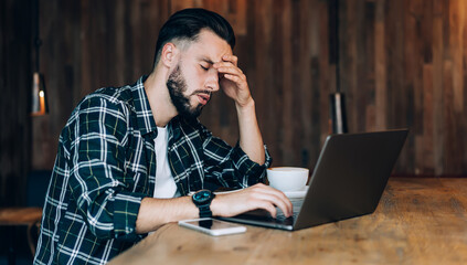 Dissatisfied bearded man during work on laptop in wooden cafe