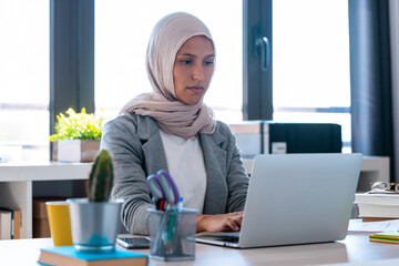 Confident young arabic business woman wearing hijab while working with laptop sitting in the office.