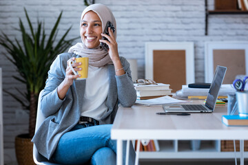 Smiling young muslim business woman wearing hijab talking with mobile phone sitting in the office.