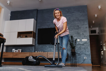 Worthy tidy smiling housewife using vacuum cleaner to vacuum dust on carpet in living room.