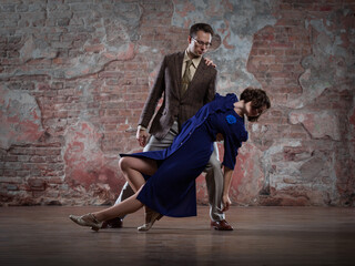 a young couple - a man in a suit and a woman in a dark-blue dress - dancing against an old brick wall