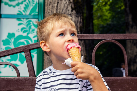 A Boy Sits On A Bench, Eats Ice Cream And Enjoys The Moment Of A Spring Or Summer Day. Positive Lifestyle And Happy Childhood Concept.