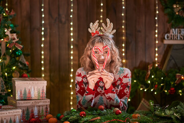 Woman dressed in christmas design sweater, deer horn hoop, handmade christmas wreath on table for holiday, making faces, have fun, posing with christmas candy. Christmas decoration and composition.