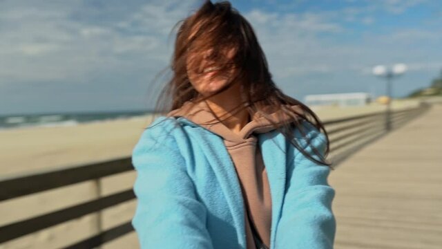 Young woman whirl around camera and happily laughing. POV holding hands with pretty woman in sky blue coat at beach with nobody in fall season. Follow me concept