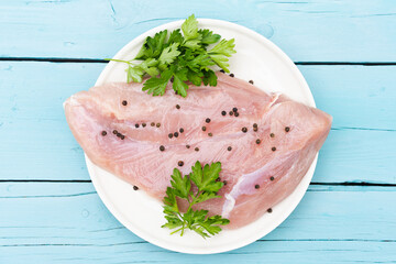 raw Turkey breast fillet on a plate, with black pepper peas and sprigs of fresh parsley, stands on a blue wooden table