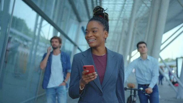 Adorable beautiful afro-american mixed race woman in business style using cellphone rejoicing message from boyfriend smiling walking near airport traveling alone.