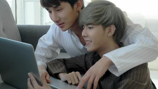 Relax and romantic LGBT couple in daily-life concept. Handheld, medium shot of southeast Asian men in living room, using laptop together, smiling, laughing, making eye contact like a loving gay couple