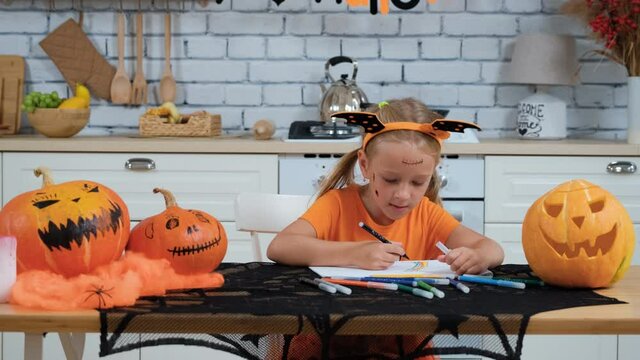 Cute Little Girl in Orange Dress Sitting at the Table and Drawing in Decorated Kitchen in Halloween. Holidays and Halloween Decorations Concept