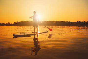 Silhouette of man paddling on paddle board at sunset. Water sport near the beach on sunset