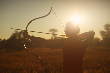 Young Caucasian female archer shooting with a bow in a field at sunset. - obrazy, fototapety, plakaty
