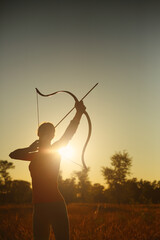 Young Caucasian female archer shooting with a bow in a field at sunset.