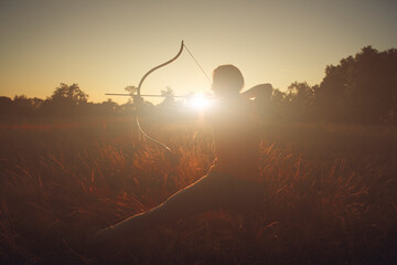 Young Caucasian female archer shooting with a bow in a field at sunset.	