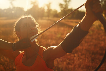 Young Caucasian female archer shooting with a bow in a field at sunset.	