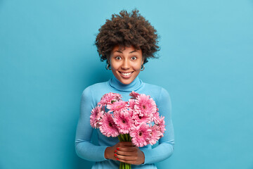 Happy joyous woman with cheerful expression, gets beautiful gerbera flowers as congratulation on birthday and wears casual blue turtleneck. Positive attractive lady likes spring nice bouquet