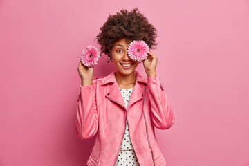 Happy dark skinned young woman covers eyes with rosy gerbera flowers, wears fashionable jacket and stands indoor. Positive professional female florist going to make bouquet for special event