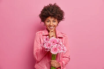 Indoor shot of pleasant looking cheerful curly woman has natural beauty, holds bouquet of flowers and going to congratulate friend on special occasion. Glad dark skinned lady with gerbera daisy