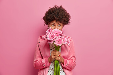 Studio shot of good looking curly haired woman smells flowers, enjoys pleasant odor and stands in...