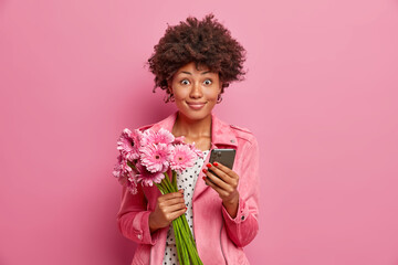 Pleased Afro American woman holds mobile phone, gets flower delivery and celebrates womens day. Smiling tender ethnic lady holds nice bouquet of gerbera daisy, surfs internet on modern device.