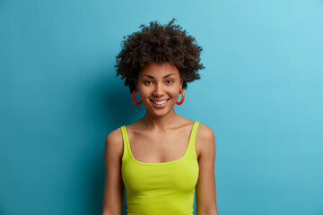 Portrait of curly haired beautiful woman with dark skin feels joyful and looks directly at camera,...