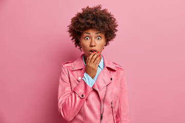 Photo of impressed curly haired adult woman keeps mouth opened and holds breath, wears stylish outfit. Shocked ethnic African American lady stares bugged eyes, stands against pink background