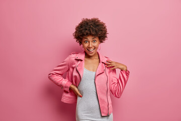 Happy surprised curly haired woman in stylish dress and jacket looks gladully at camera has joy keeps hand on waist, isolated on pink background. Delighted Afro American woman in fashionable outfit