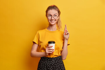 Smiling beautiful European girl raises hand and crosses fingers for luck, holds disposable cup of coffee. Happy Caucasian schoolgirl believes in receiving good mark for test, poses over yellow wall