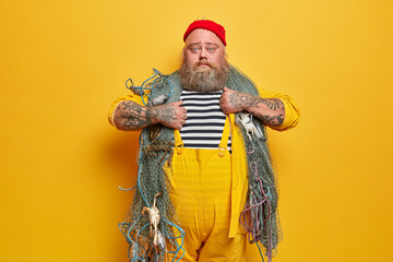 Serious plump bearded experienced man sailor poses with fishing gears, wears striped sailor shirt...