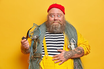 Plump surprised boatswain or sailor has tattooed arms, holds smoking pipe and goes fishing at sea. Shocked sailor focused at camera, wears red hat yellow overalls. Sea adventure hobby concept - obrazy, fototapety, plakaty