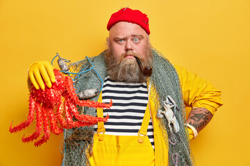 Horizontal shot of self confident sailor has thick beard and holds big red octopus caught during...