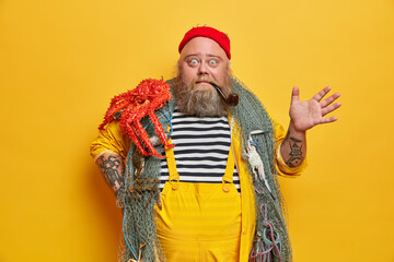 Surprised emotional seafarer smokes pipe and raised hand, stares bugged eyes at camera. Bearded boatswain poses with sea creature against vivid yellow background. Fisherman caught big red octopus