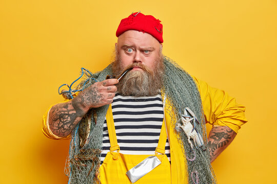 Close up shot of serious bearded sailor smokes pipe and looks unhappily at camera, poses with fishing net against yellow studio wall. Angry tattooed fisherman in overalls spends free time at sea