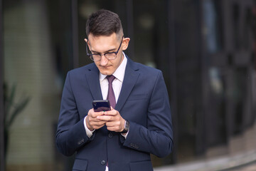 Serious businessman in formal suit, tie and glasses looking at cell mobile phone, young man typing a message at social media on his smartphone