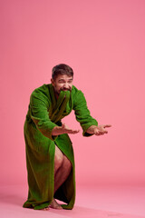 An emotional man in a green robe in full growth on a pink background gestures with his hands to the model