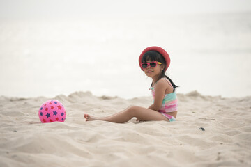 Happy asian child girl playing on white sand at the beach in summer. Asia kid girl with sunglasses and pink hat joyful on sand beach in thailand. people travel holiday concept