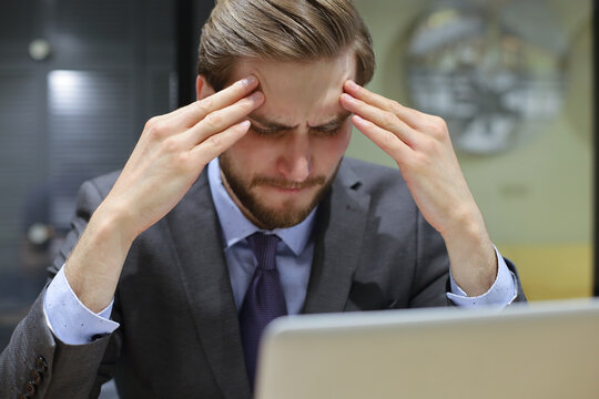 Image of tired young business man in suit keeping face in hands while sitting in the office.