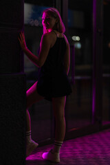 Obraz na płótnie Canvas Club style photo of girl in a black dress, white socks and sneakers. Set is lit with violet light. Picture has dark night tone. She stands at the entrance to the building